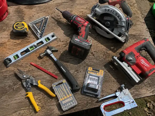 How to Build a Chicken Coop?-Beginner's Guide 13 Essential chicken coop construction tools arranged on a wooden workbench, including a tape measure, level, speed square, pencil, circular saw, jigsaw, cordless drill, hammer, drill bits, wire cutters, and staple gun