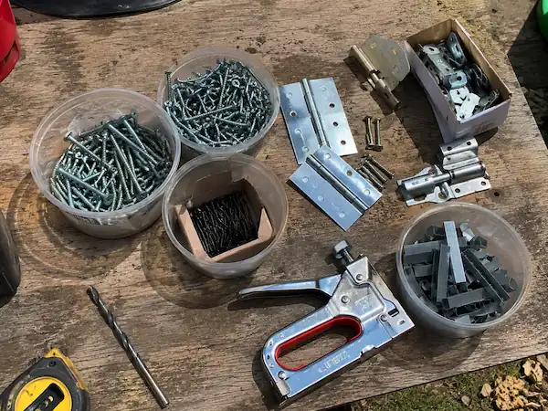 How to Build a Chicken Coop?-Beginner's Guide 12 Collection of chicken coop building hardware on a workbench, including exterior screws, nails, door hinges, latch locks, a staple gun, and fencing staples used for coop construction